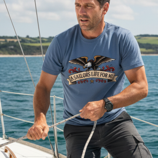 A man on a sailboat wearing a slate blue graphic t-shirt with an eagle design and the text 'A SAILORS LIFE FOR ME'.