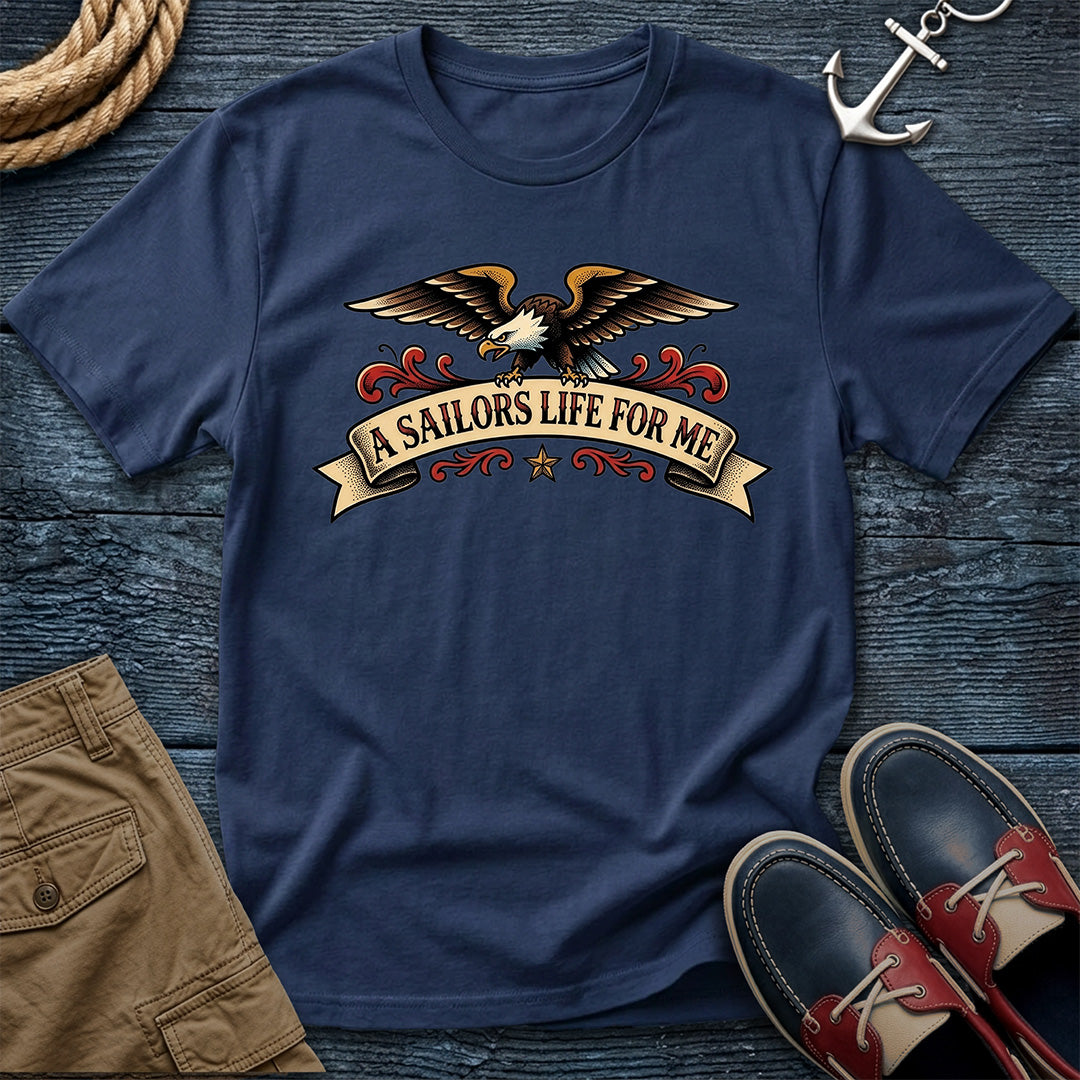 A navy blue crewneck t-shirt with a graphic of an eagle and the text 'A SAILORS LIFE FOR ME' on a banner, styled in a flat lay with brown shorts, red and navy boat shoes, a rope, and an anchor on a dark wood background.
