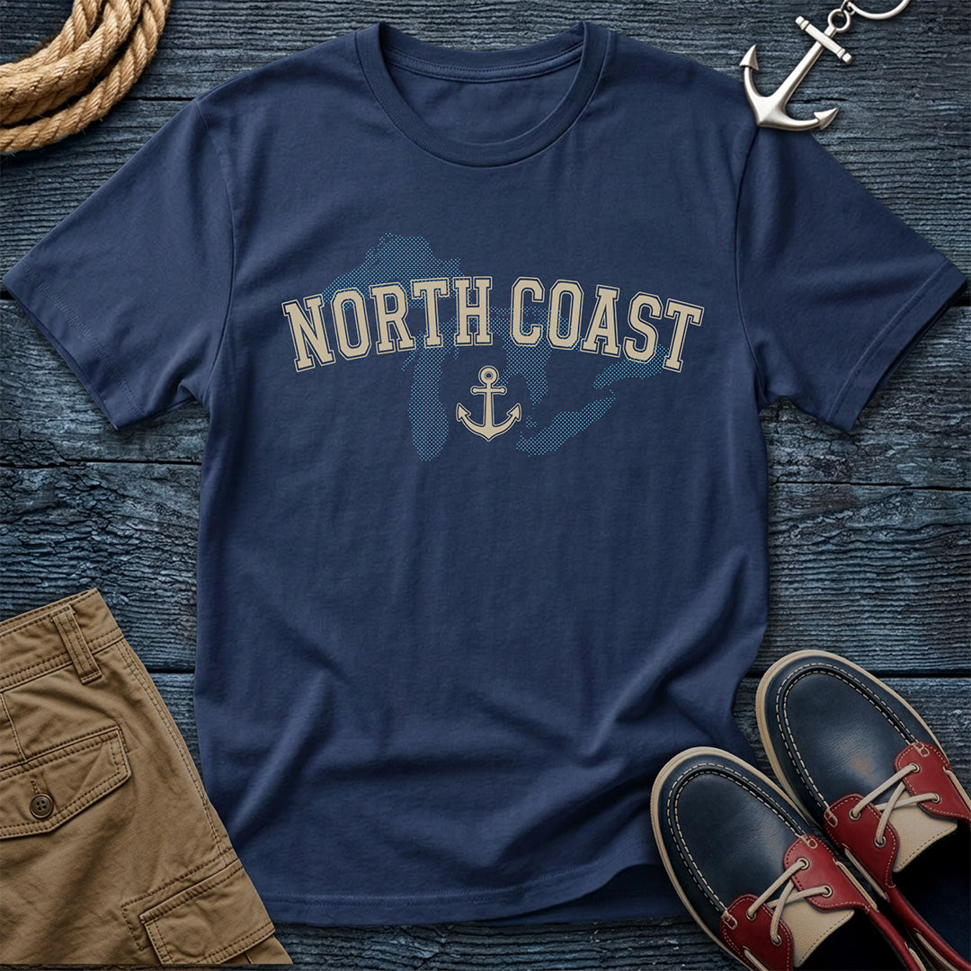 A navy blue t-shirt with 'NORTH COAST' and an anchor graphic printed on the chest, styled in a flat-lay with brown shorts, red and navy boat shoes, a rope, and an anchor on a dark wood background.