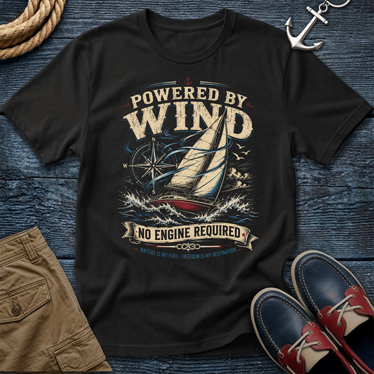 A black short-sleeve graphic T-shirt laid flat on a dark blue wood surface, surrounded by nautical props including a rope, anchor, tan shorts, and blue-and-red boat shoes. The shirt graphic shows a sailboat with the text 'POWERED BY WIND' and 'NO ENGINE REQUIRED.