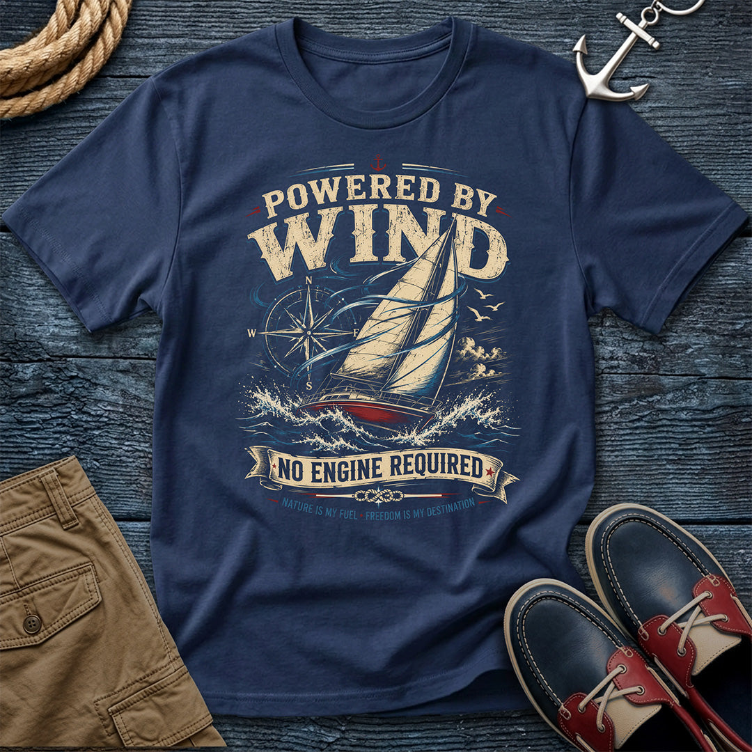 A Navy colored graphic t-shirt featuring a sailboat and the text 'POWERED BY WIND NO ENGINE REQUIRED' in a vintage nautical design. The shirt is styled in a flat-lay arrangement on a dark wood background with a rope, khaki shorts, an anchor, and red-and-navy boat shoes.