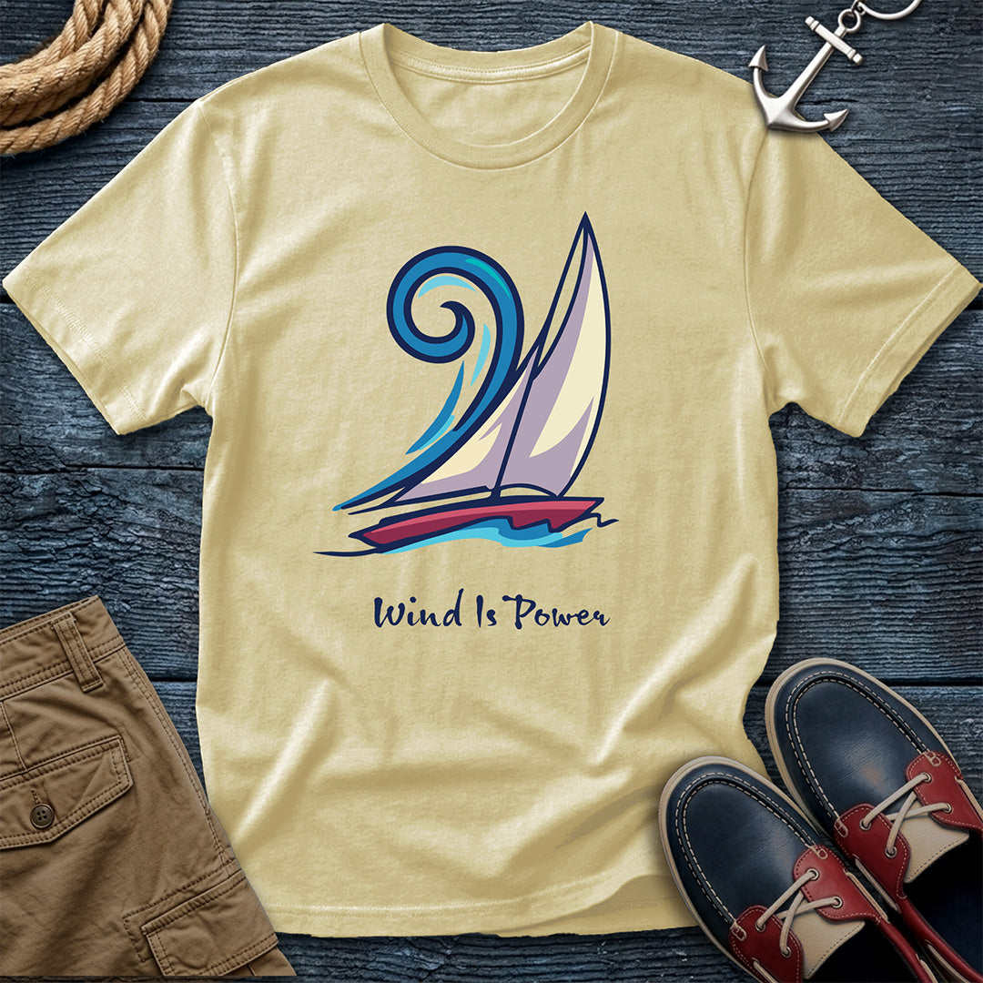 A yellow crewneck t-shirt with a graphic of a sailboat and the text 'Wind Is Power', styled in a flat lay on a dark wood background with a rope, an anchor, brown shorts, and navy shoes.