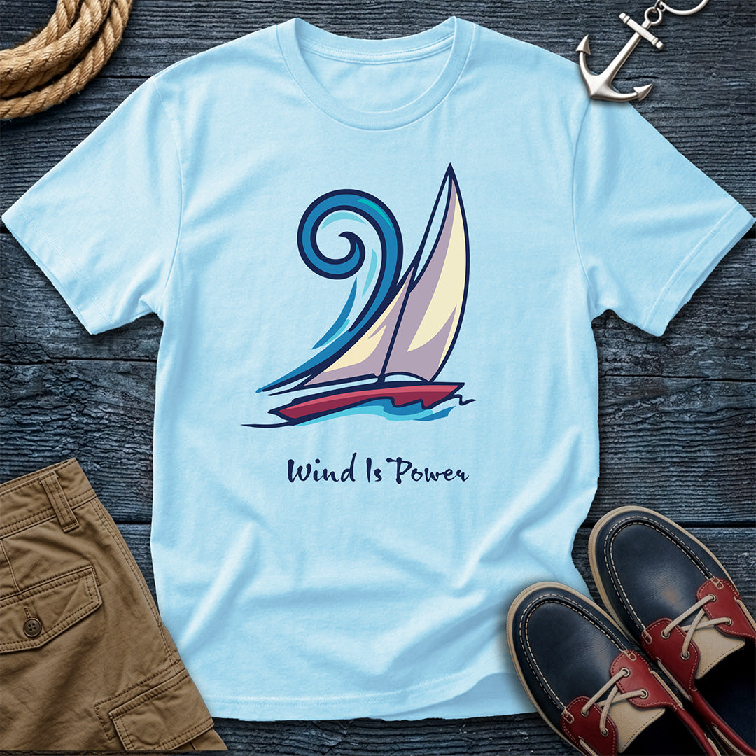 A light blue crewneck t-shirt featuring a graphic of a sailboat with the text 'Wind Is Power' underneath, styled in a flat lay with brown shorts, boat shoes, rope, and an anchor on a dark wood background.