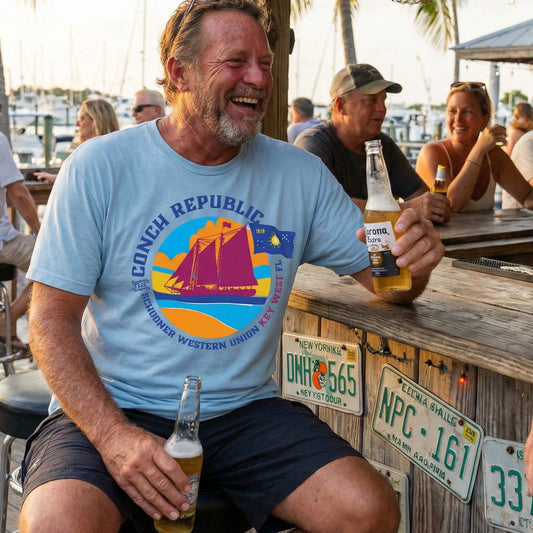 Man wearing the Conch Republic Key West Schooner Tee in Chambray at an outdoor waterfront bar, showing the colorful Schooner Western Union and Conch Republic flag graphic.