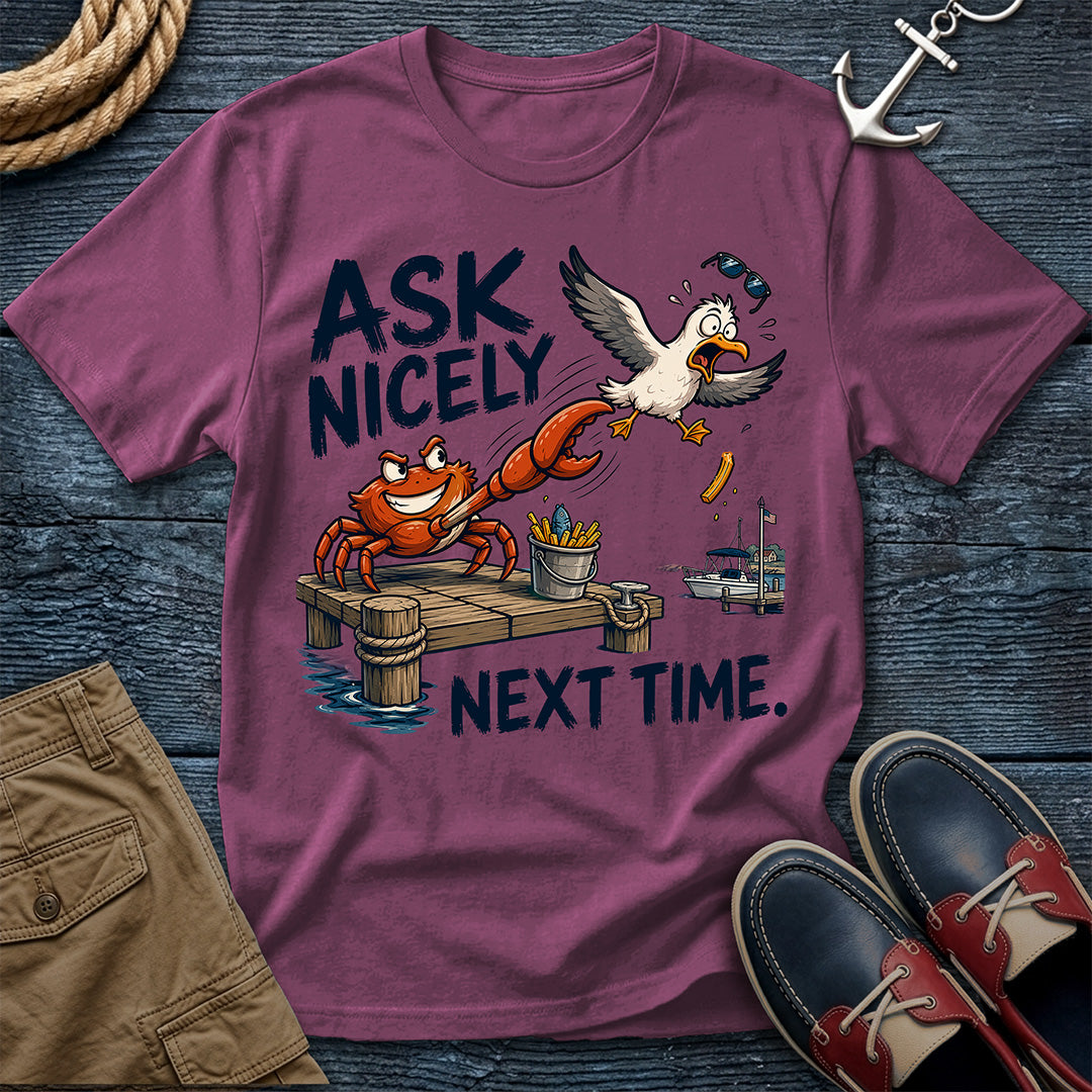 A mauve purple graphic t-shirt laid flat on a dark wood background, surrounded by nautical-themed props including a rope, anchor, shorts, and boat shoes. The t-shirt features a cartoon graphic of a crab on a dock with a seagull wearing sunglasses and the text 'ASK NICELY NEXT TIME.