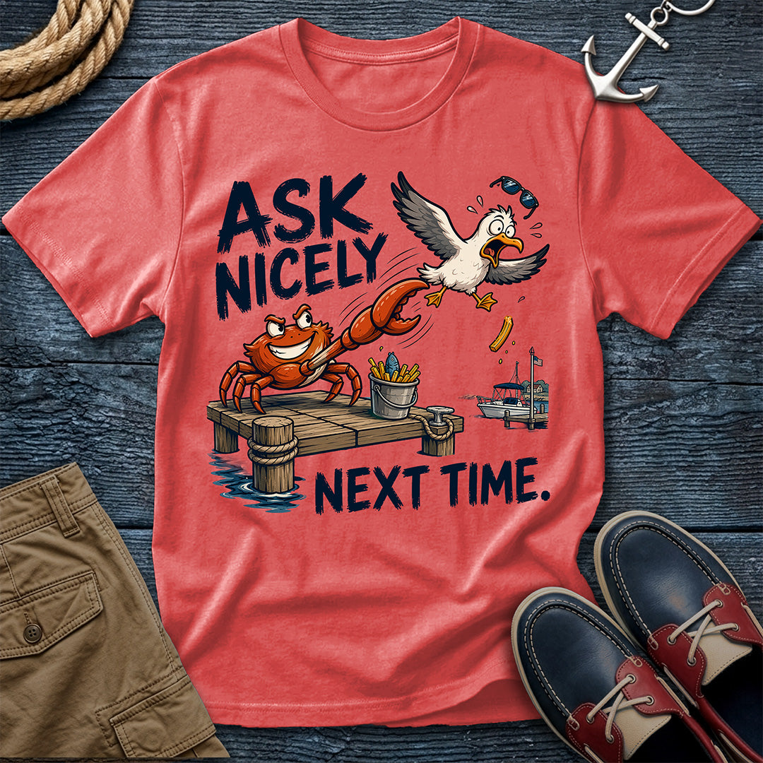 A coral red graphic t-shirt laid flat on a dark wood background, surrounded by nautical and casual props including rope, an anchor, khaki shorts, and navy-and-red boat shoes. The shirt features a cartoon crab and seagull at a dock with the text 'ASK NICELY NEXT TIME.