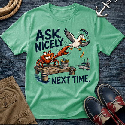 A mint green graphic t-shirt laid flat on a dark wood background, surrounded by nautical-themed props including a rope, an anchor, khaki shorts, and navy and red boat shoes. The t-shirt features a cartoon graphic of a crab on a dock trying to flirt with a seagull while the text reads 'ASK NICELY NEXT TIME.