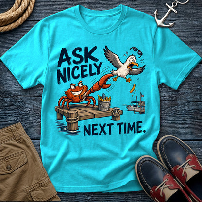 A turquoise graphic T-shirt laid flat on a dark wood surface, featuring a cartoon crab and seagull scene with the text 'ASK NICELY NEXT TIME.' Surrounding props include rope, an anchor, tan shorts, and navy-and-red boat shoes.