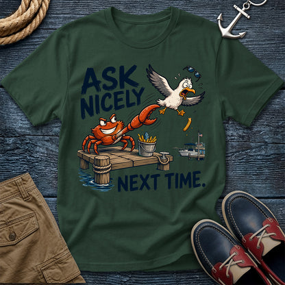 A dark green graphic t-shirt laid flat on a dark blue wooden surface, surrounded by nautical-themed props including a rope, an anchor, khaki shorts, and blue-and-red boat shoes. The t-shirt features a cartoon graphic of a red crab on a dock with the text 'ASK NICELY NEXT TIME.