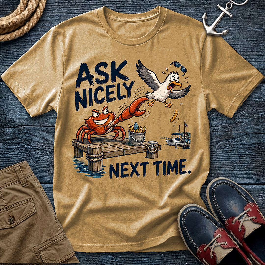 A tan graphic t-shirt laid flat on a dark blue wood background, featuring a cartoon crab and seagull with the text 'ASK NICELY NEXT TIME.' Surrounding props include rope, an anchor, khaki shorts, and navy-and-red boat shoes.