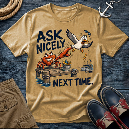 A tan graphic t-shirt laid flat on a dark blue wood background, featuring a cartoon crab and seagull with the text 'ASK NICELY NEXT TIME.' Surrounding props include rope, an anchor, khaki shorts, and navy-and-red boat shoes.
