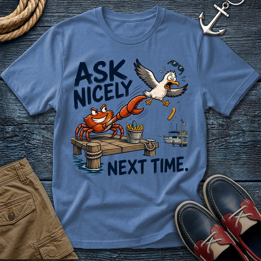A blue graphic t-shirt laid flat on a dark wood background, featuring a cartoon crab, seagull, dock, and the text 'ASK NICELY NEXT TIME.' Surrounding props include rope, an anchor, khaki shorts, and red-and-black boat shoes.