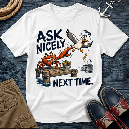A white graphic t-shirt laid flat on a dark blue wood background, featuring a cartoon crab and seagull scene with the text 'ASK NICELY NEXT TIME.' Brown shorts, navy and red boat shoes, rope, and an anchor are styled around the shirt.