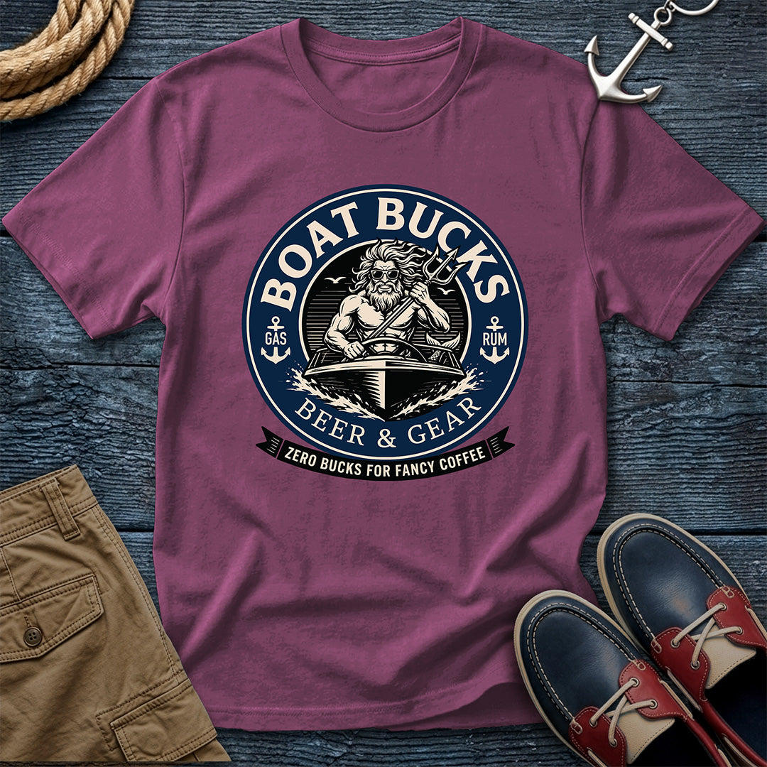 A berry short-sleeve graphic T-shirt laid flat on a dark wood surface, surrounded by nautical and casual styling props including rope, an anchor, tan shorts, and navy-and-red boat shoes. The shirt features a large circular front print reading “BOAT BUCKS BEER & GEAR” with an illustrated sailor and smaller text “ZERO BUCKS FOR FANCY COFFEE.