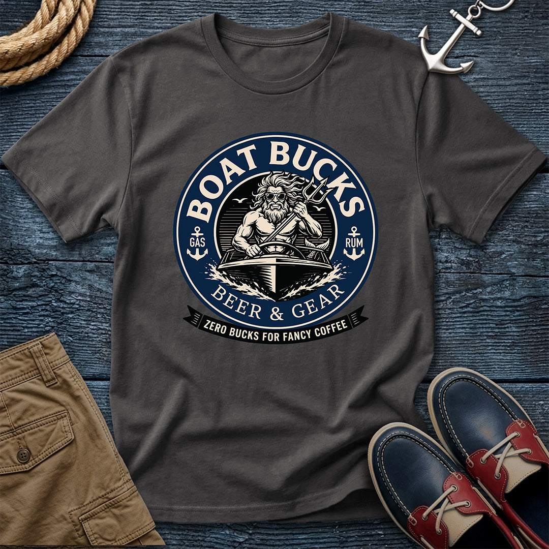 A dark charcoal gray graphic t-shirt laid flat on a dark blue wooden surface, surrounded by nautical-themed props including a rope, an anchor, khaki shorts, and red-and-navy boat shoes. The shirt features a circular graphic with a sailor and the text 'BOAT BUCKS BEER & GEAR'.