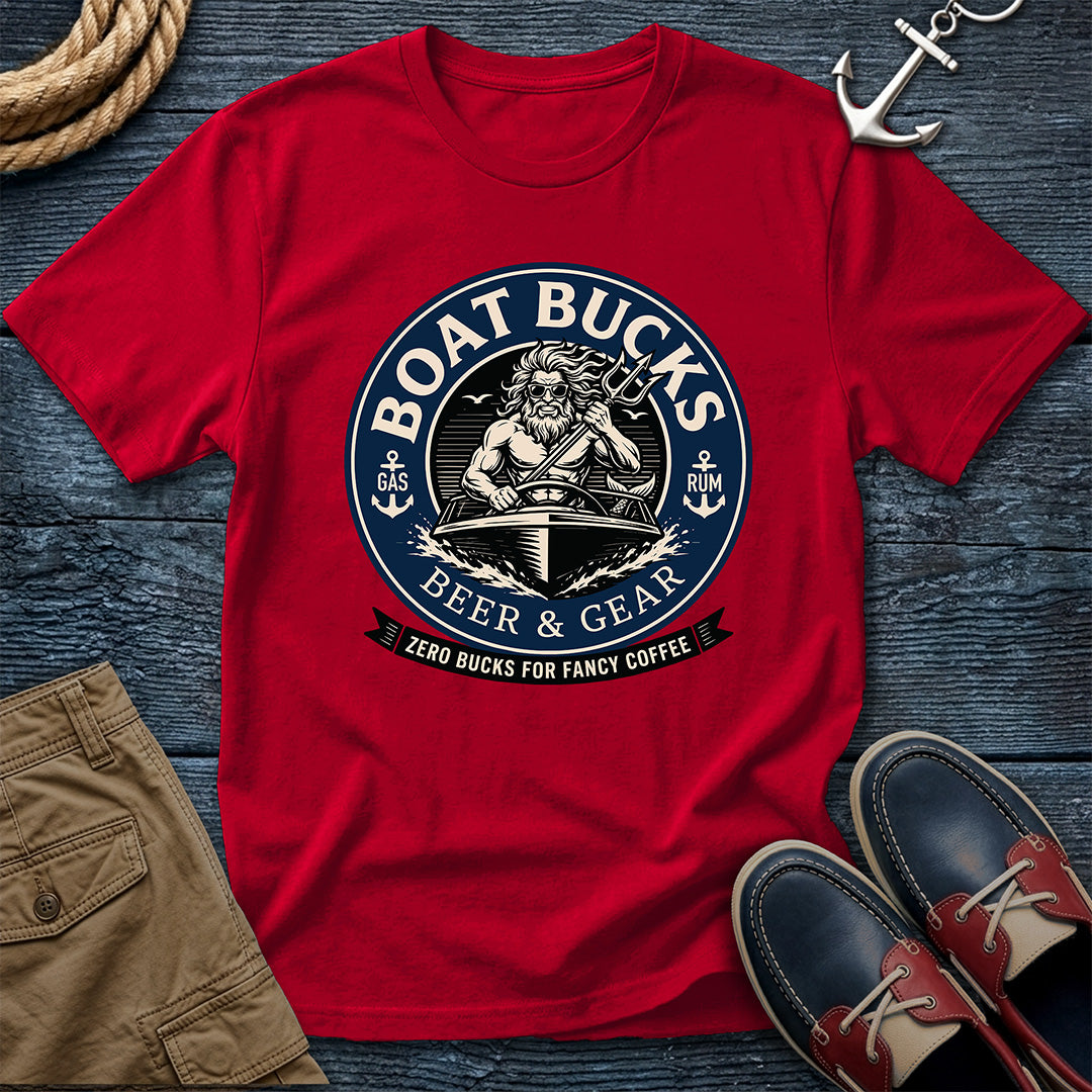 A red short-sleeve graphic T-shirt laid flat on a dark wood surface, styled with a rope, anchor, khaki shorts, and navy-and-red boat shoes. The shirt has a large circular nautical-style print reading “BOAT BUCKS BEER & GEAR” with an anchor, ship wheel, and banner reading “ZERO BUCKS FOR FANCY COFFEE.