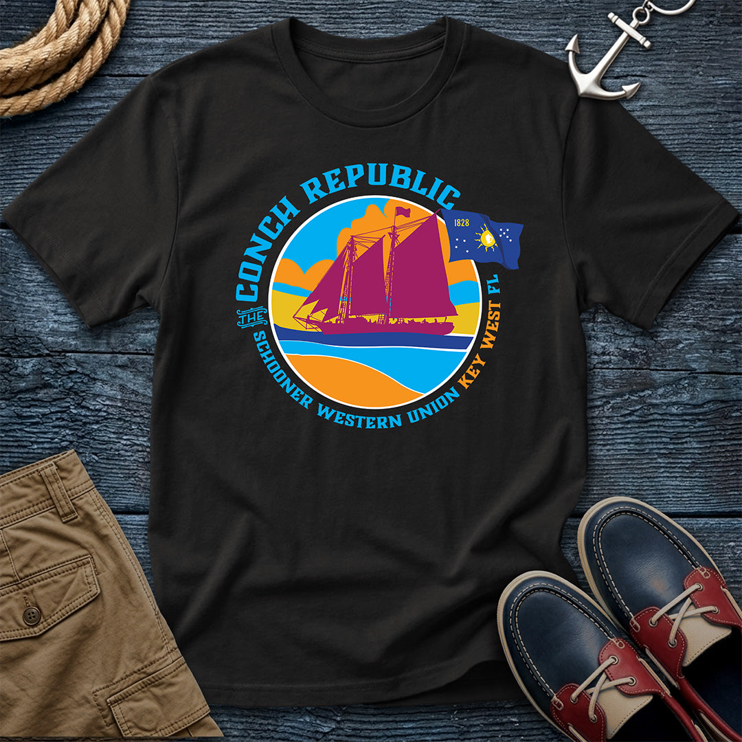 Conch Republic Key West Schooner Tee in Black, Comfort Colors 1717, featuring a magenta schooner, blue Conch Republic flag, and orange sunset graphic, laid flat beside nautical accessories.