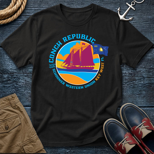 Conch Republic Key West Schooner Tee in Black, Comfort Colors 1717, featuring a magenta schooner, blue Conch Republic flag, and orange sunset graphic, laid flat beside nautical accessories.
