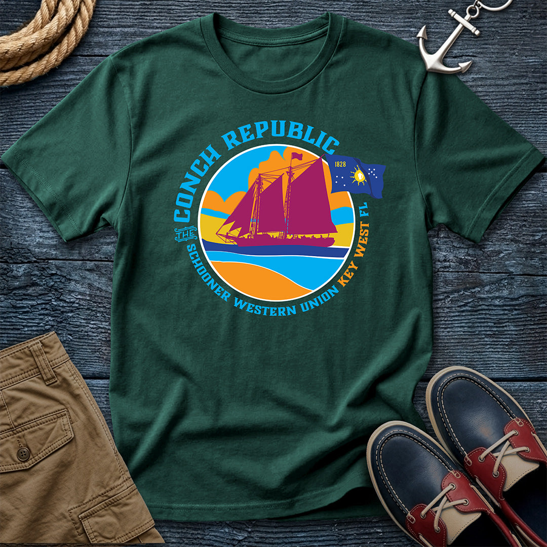 Conch Republic Key West Schooner Tee in Blue Spruce, Comfort Colors 1717, featuring a magenta schooner, blue Conch Republic flag, and orange sunset graphic, laid flat beside nautical accessories.