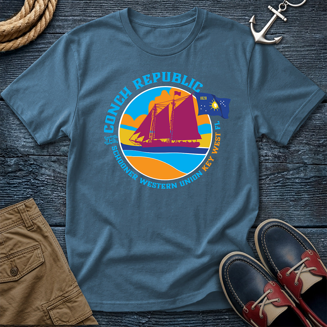 Conch Republic Key West Schooner Tee in Denim, Comfort Colors 1717, featuring a magenta schooner, blue Conch Republic flag, and orange sunset graphic, laid flat beside nautical accessories.