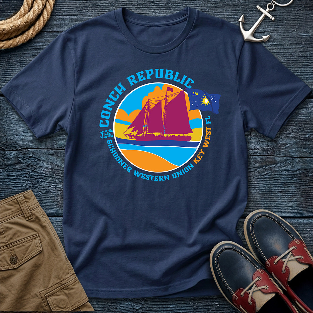 Conch Republic Key West Schooner Tee in Navy, Comfort Colors 1717, featuring a magenta schooner, blue Conch Republic flag, and orange sunset graphic, laid flat beside nautical accessories.