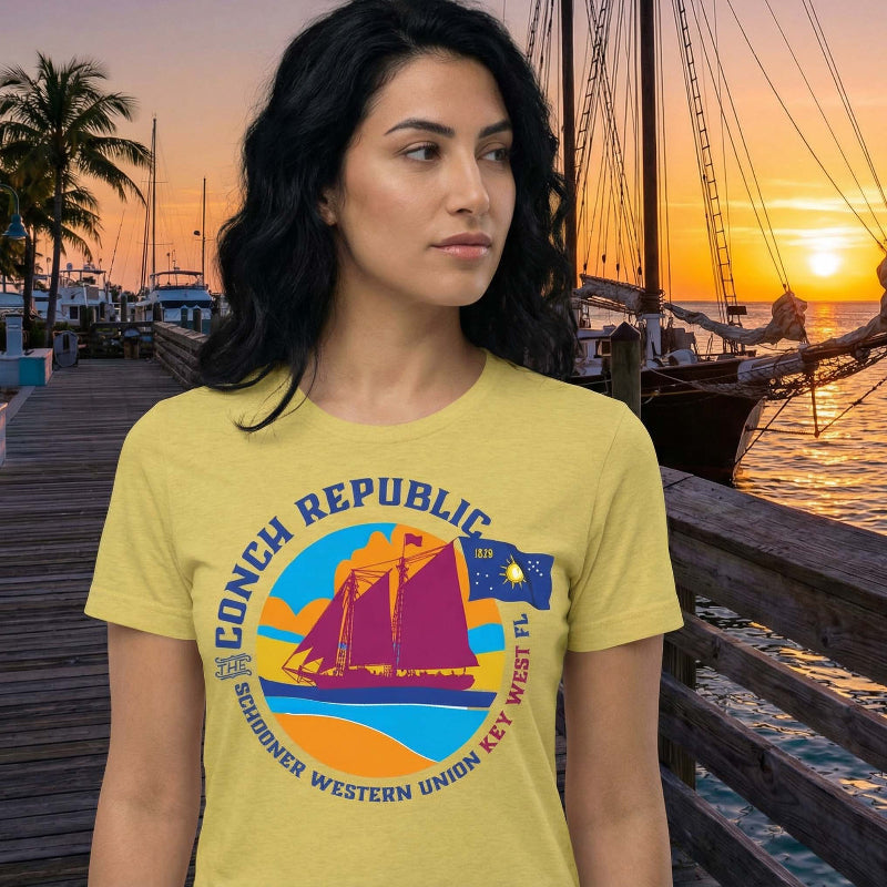 Woman wearing the Conch Republic Key West Schooner Tee in Butter beside a docked schooner at sunset, showing the colorful Western Union schooner and Conch Republic flag graphic.