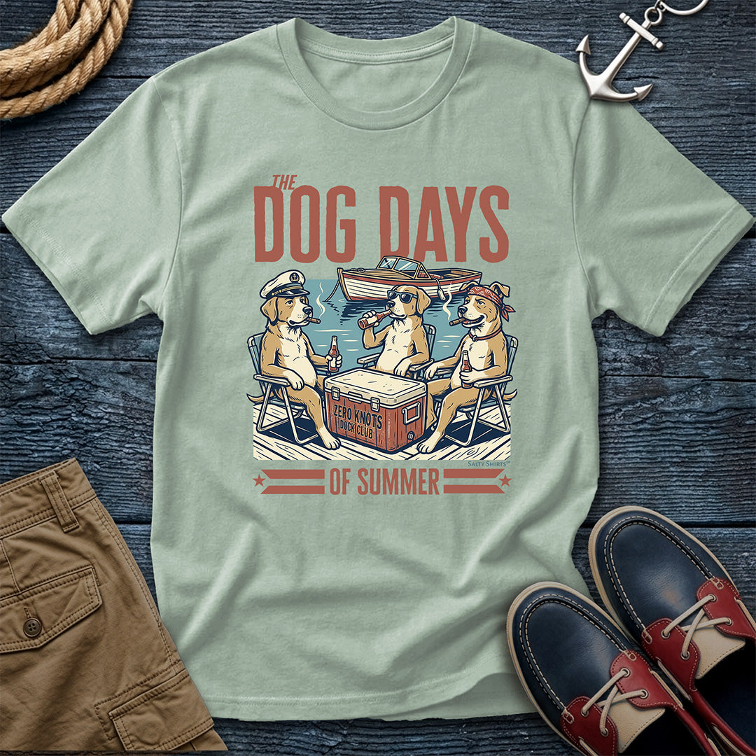A light sage green t-shirt with a retro graphic of three dogs at a summer dock, styled in a flat-lay with brown shorts, a rope, an anchor, and boat shoes on a dark wood background.