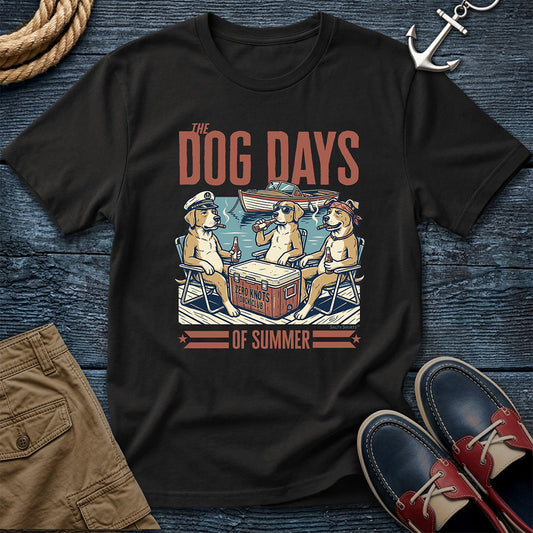 A black graphic t-shirt featuring a retro-style illustration of two dogs relaxing in chairs by a cooler with the text 'THE DOG DAYS OF SUMMER'. The shirt is presented in a flat-lay arrangement on a dark blue wooden background, styled with a rope, an anchor, khaki shorts, and red-and-navy boat shoes.