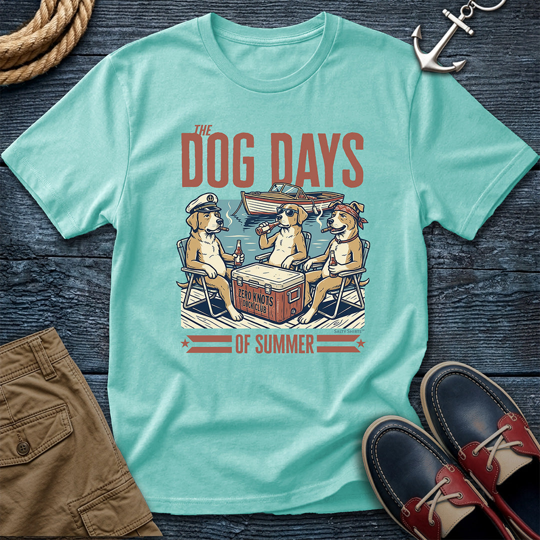 A Tiffany mint green graphic t-shirt laid flat on a dark wood background, styled with brown shorts, a rope, an anchor, and red-and-black boat shoes. The t-shirt features a retro-style illustration of three dogs sitting in chairs by a cooler under the text 'THE DOG DAYS OF SUMMER'.