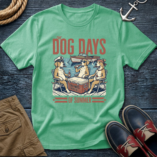 A mint green graphic t-shirt laid flat on a dark wood background, featuring a retro-style illustration of three dogs sitting in beach chairs around a cooler with the text 'THE DOG DAYS OF SUMMER'. The shirt is styled with brown shorts, navy and red boat shoes, a rope, and an anchor.