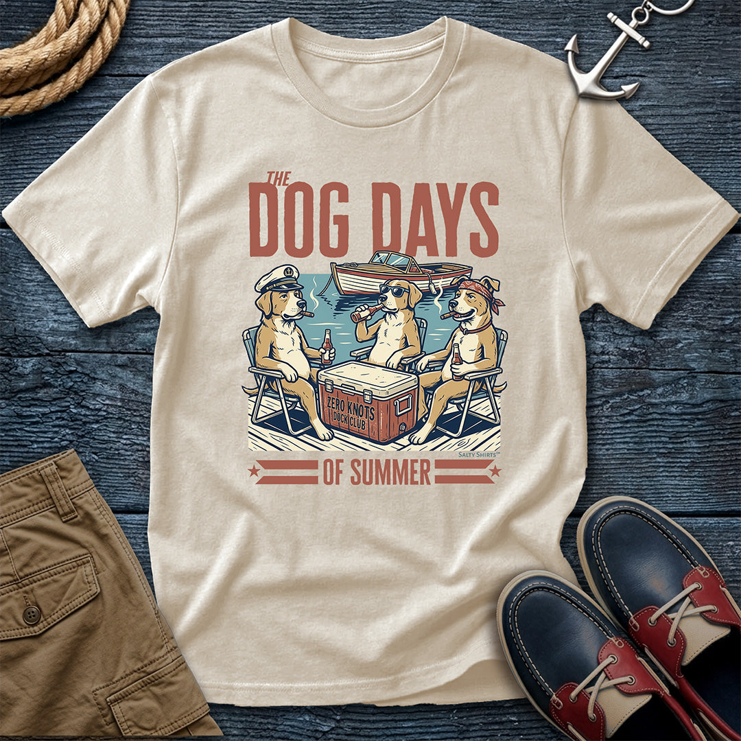 An Ivory graphic t-shirt laid flat on a dark blue wooden surface, featuring a vintage-style illustration of three dogs sitting in chairs around a cooler with the text 'THE DOG DAYS OF SUMMER'. The shirt is styled with brown shorts, a rope, an anchor, and red-and-navy shoes.