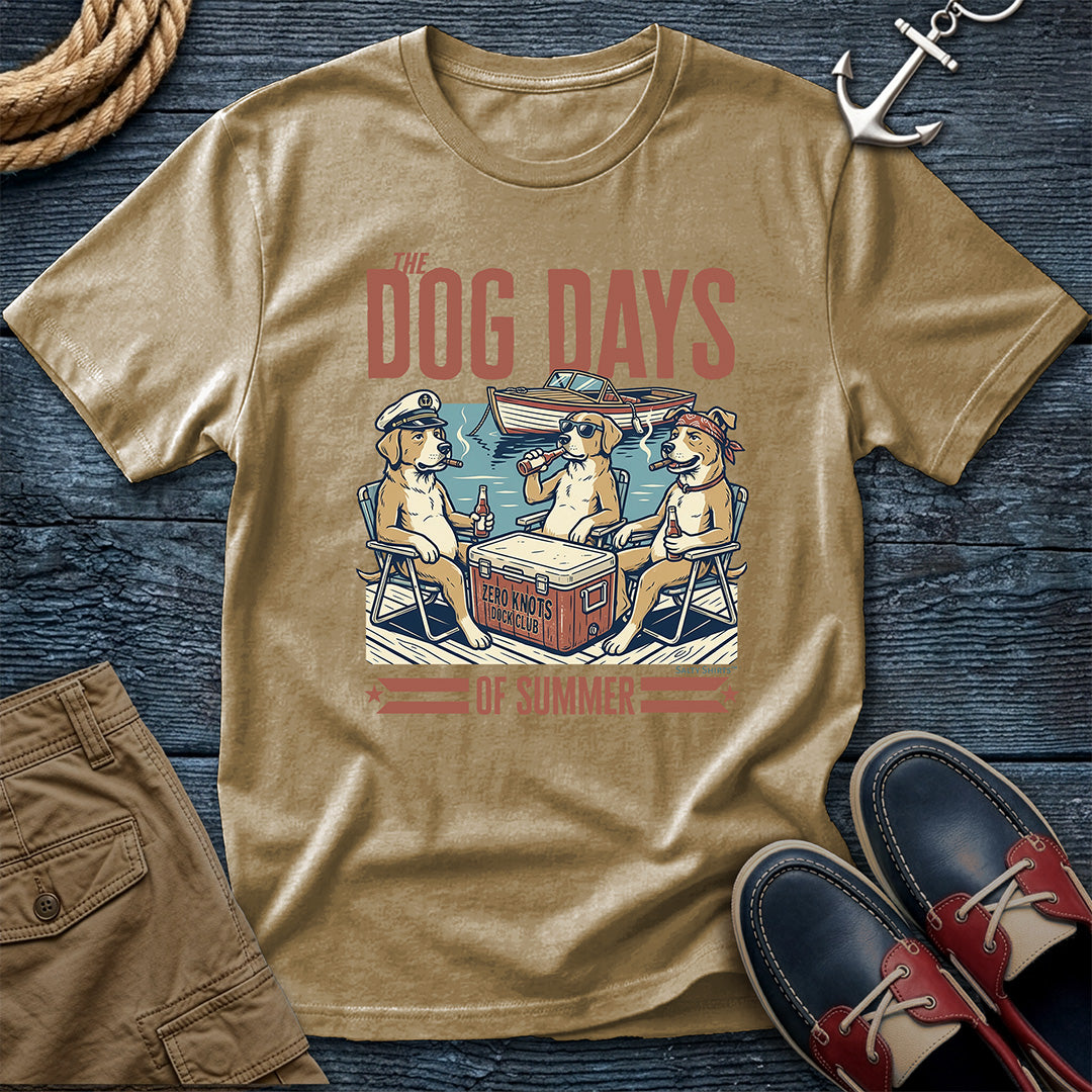 A Khaki graphic t-shirt laid flat on a dark blue wood background, styled with a rope, anchor, khaki shorts, and navy-and-red boat shoes. The shirt features a retro-style print of three dogs sitting in camp chairs by a cooler with the text 'THE DOG DAYS OF SUMMER'.