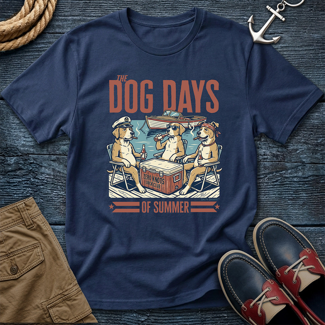A navy blue graphic t-shirt laid flat on a dark wood background, featuring an illustration of three dogs relaxing at a cooler with the text 'THE DOG DAYS OF SUMMER'. The shirt is styled with brown shorts, red and navy boat shoes, a rope, and an anchor.