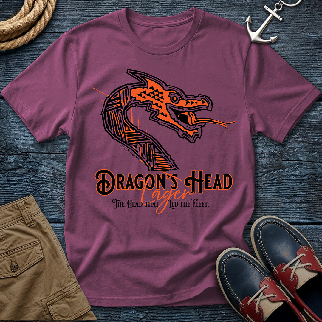 A mauve purple t-shirt laid flat on a dark wood background, featuring a graphic of a stylized orange and black dragon head with the text 'DRAGON'S HEAD Lager' and 'The Head That Led The Fleet'. The shirt is styled with a rope, khaki shorts, a silver anchor, and navy and red boat shoes.