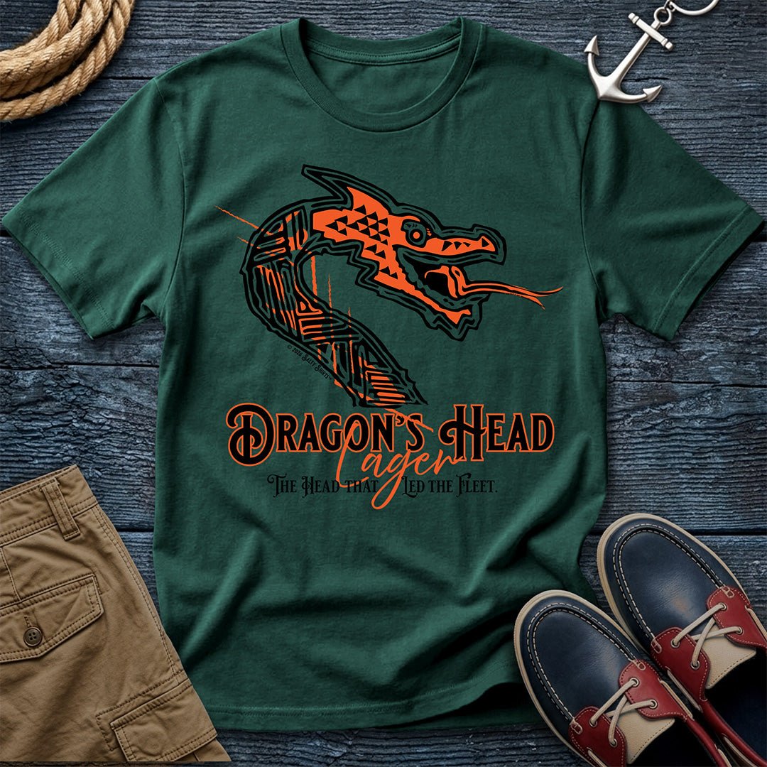 A dark green crewneck t-shirt featuring a stylized orange and black dragon head graphic with the text 'DRAGON'S HEAD Lager' and 'The Head of the Fleet' printed on the front. The shirt is presented in a flat-lay arrangement on a dark wood background, styled with a rope, khaki shorts, an anchor, and boat shoes.