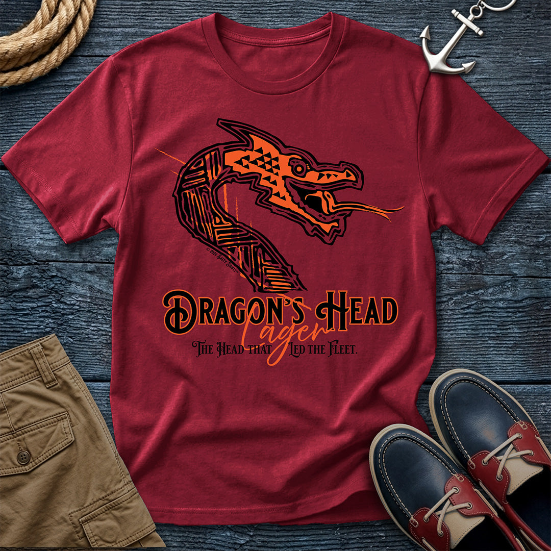 A Chili short-sleeve graphic T-shirt laid flat on a dark wood surface, styled with rope, an anchor, tan shorts, and navy-and-red boat shoes. The shirt features a large orange and black dragon head illustration and the text 'DRAGON'S HEAD Lager' with smaller text below.