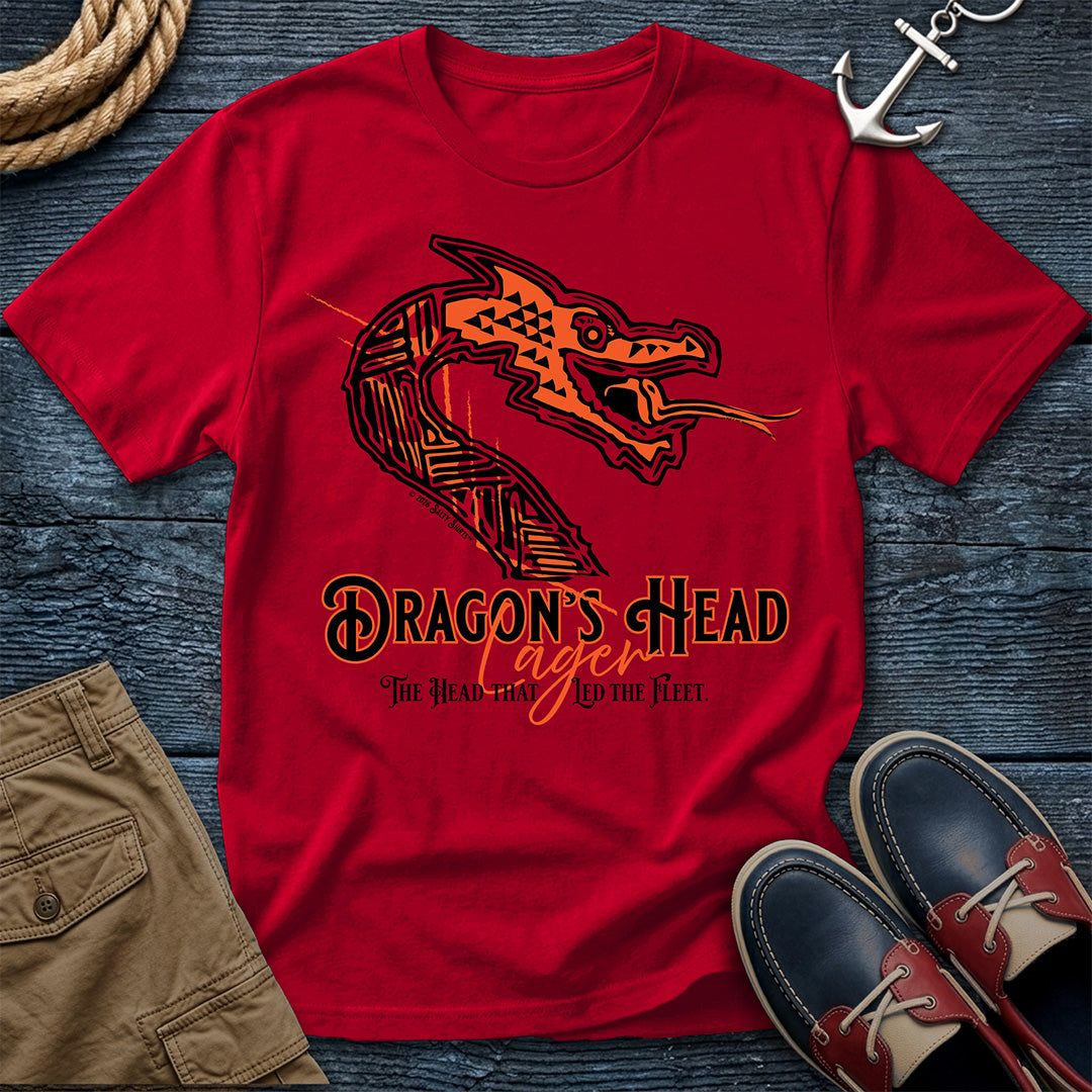 A red short-sleeve graphic T-shirt laid flat on a dark wood surface, styled with tan shorts, navy shoes, rope, and an anchor. The shirt features a large illustrated dragon head in orange and black with the text 'Dragon's Head' and smaller text beneath.