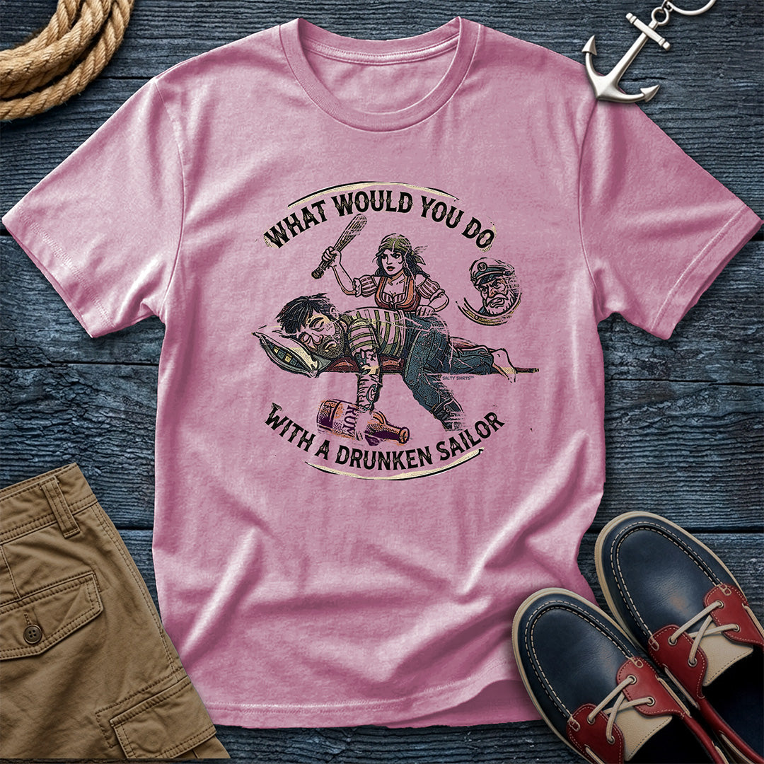 Drunken Sailor Shanty Tee in Blossom: flat lay of a Comfort Colors 1717 tee on dark weathered wood with rope, an anchor keychain, khaki pants, and boat shoes. Distressed woodcut print shows a woman with a club swinging at a passed-out drunken sailor holding a rum bottle, framed by the text "What Would You Do With a Drunken Sailor."