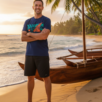 A man stands on a sandy beach with the ocean and palm trees behind him, wearing a navy blue graphic of Koa Sailboat t-shirt and black shorts. A wooden outrigger canoe is visible on the right side of the frame.