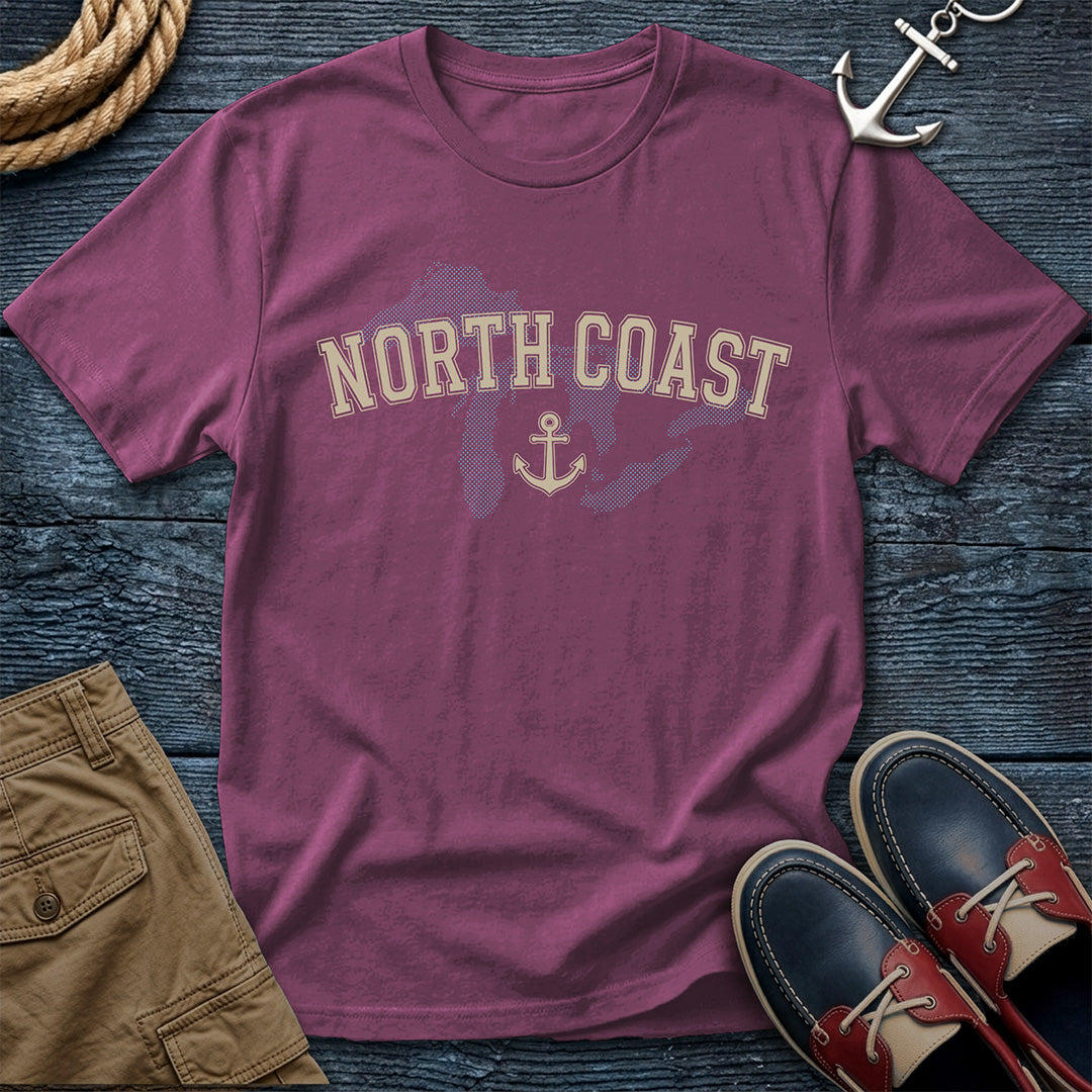 A mauve-purple crewneck t-shirt with 'NORTH COAST' and an anchor graphic printed on the front, styled in a flat-lay with boat shoes, shorts, rope, and an anchor on a dark wood background.