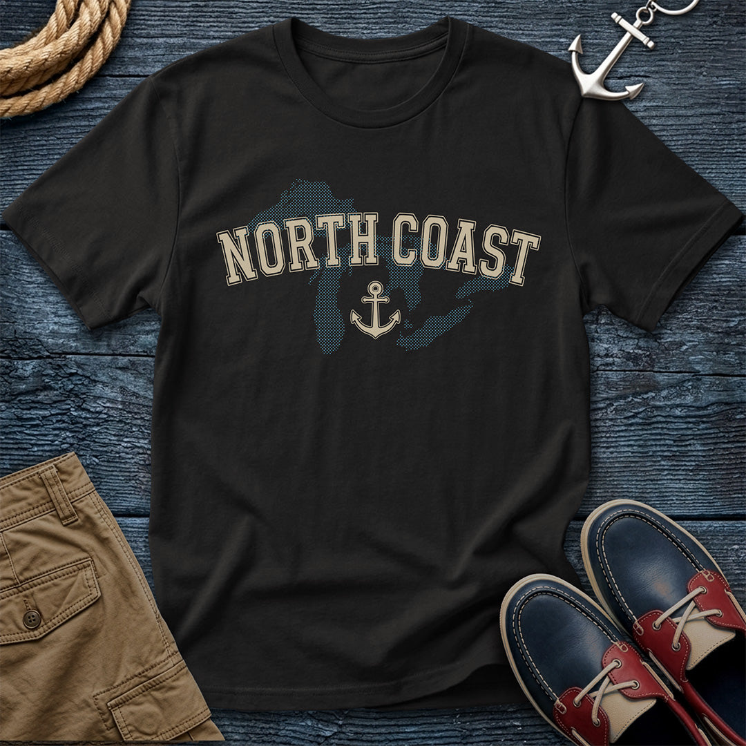 A black short-sleeve graphic T-shirt laid flat on a dark blue wood surface, styled with a rope coil, silver anchor keychain, tan shorts, and red-and-navy boat shoes. The shirt has a distressed cream 'NORTH COAST' text across the chest with a small anchor graphic and a muted blue map silhouette behind it.