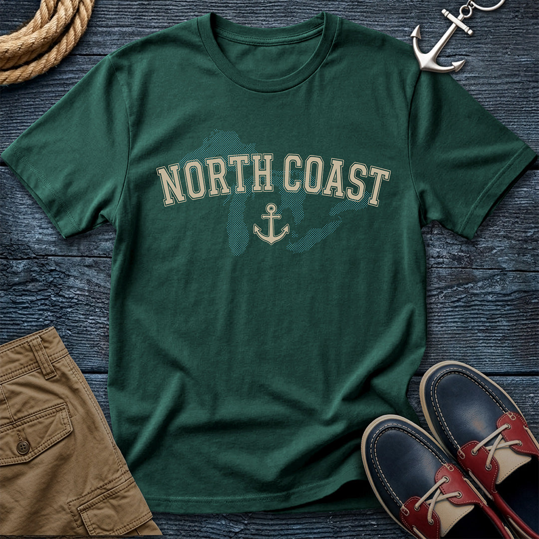 A dark green short-sleeve t-shirt with 'NORTH COAST' and an anchor graphic printed on the front, styled in a flat lay with brown shorts, red and navy boat shoes, a rope, and an anchor on a dark wood background.