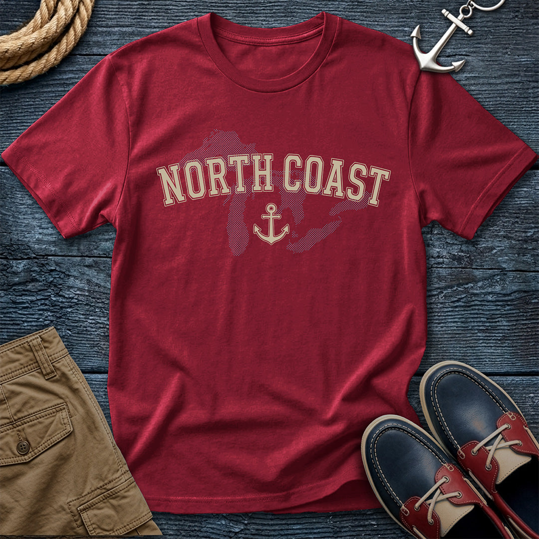 A red short-sleeve graphic T-shirt laid flat on a dark blue wood surface, with 'NORTH COAST' and an anchor printed across the chest. Surrounding props include a rope coil, khaki shorts, a silver anchor ornament, and navy-and-red boat shoes.