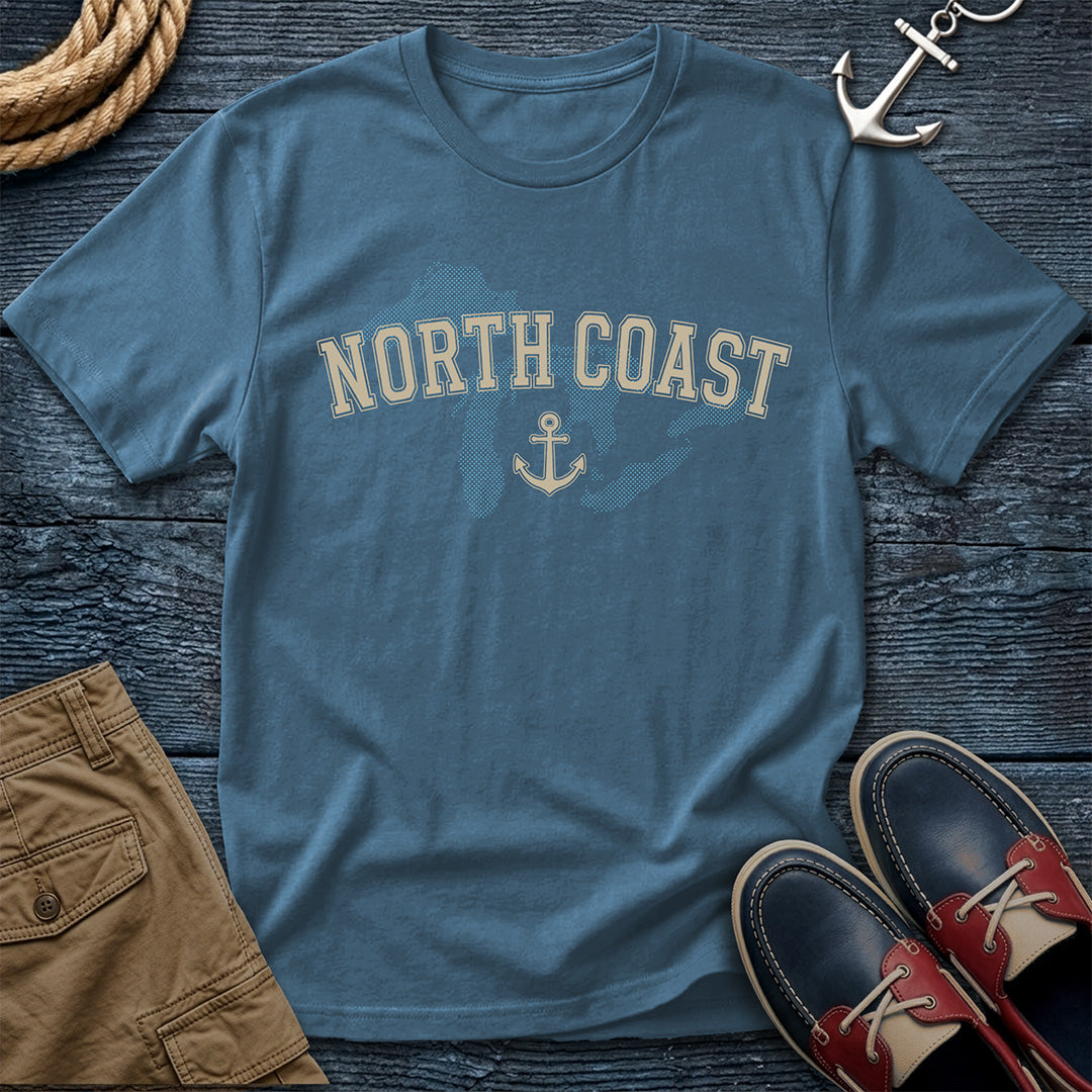 A slate blue crewneck t-shirt with 'NORTH COAST' and an anchor graphic printed on the front, styled in a flat lay with brown shorts, red and navy boat shoes, a rope coil, and an anchor charm on a dark wood background.