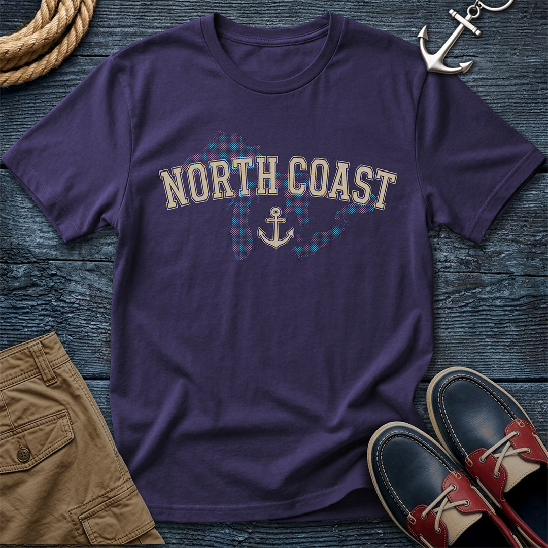 A purple short-sleeve t-shirt with a distressed cream 'NORTH COAST' text and anchor graphic over a blue silhouette map, styled in a flat lay with khaki shorts, red-and-black boat shoes, rope, and an anchor on a dark wood background.