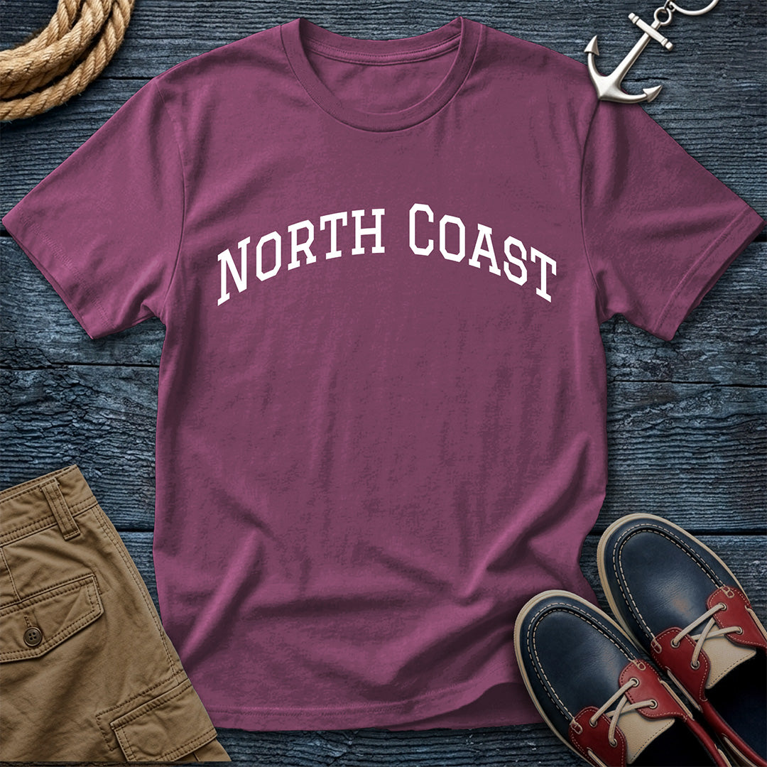A maroon short-sleeve t-shirt with 'NORTH COAST' printed in white arched letters across the chest, styled in a flat lay on a dark wood background with a rope, silver anchor, tan shorts, and red-and-navy boat shoes.