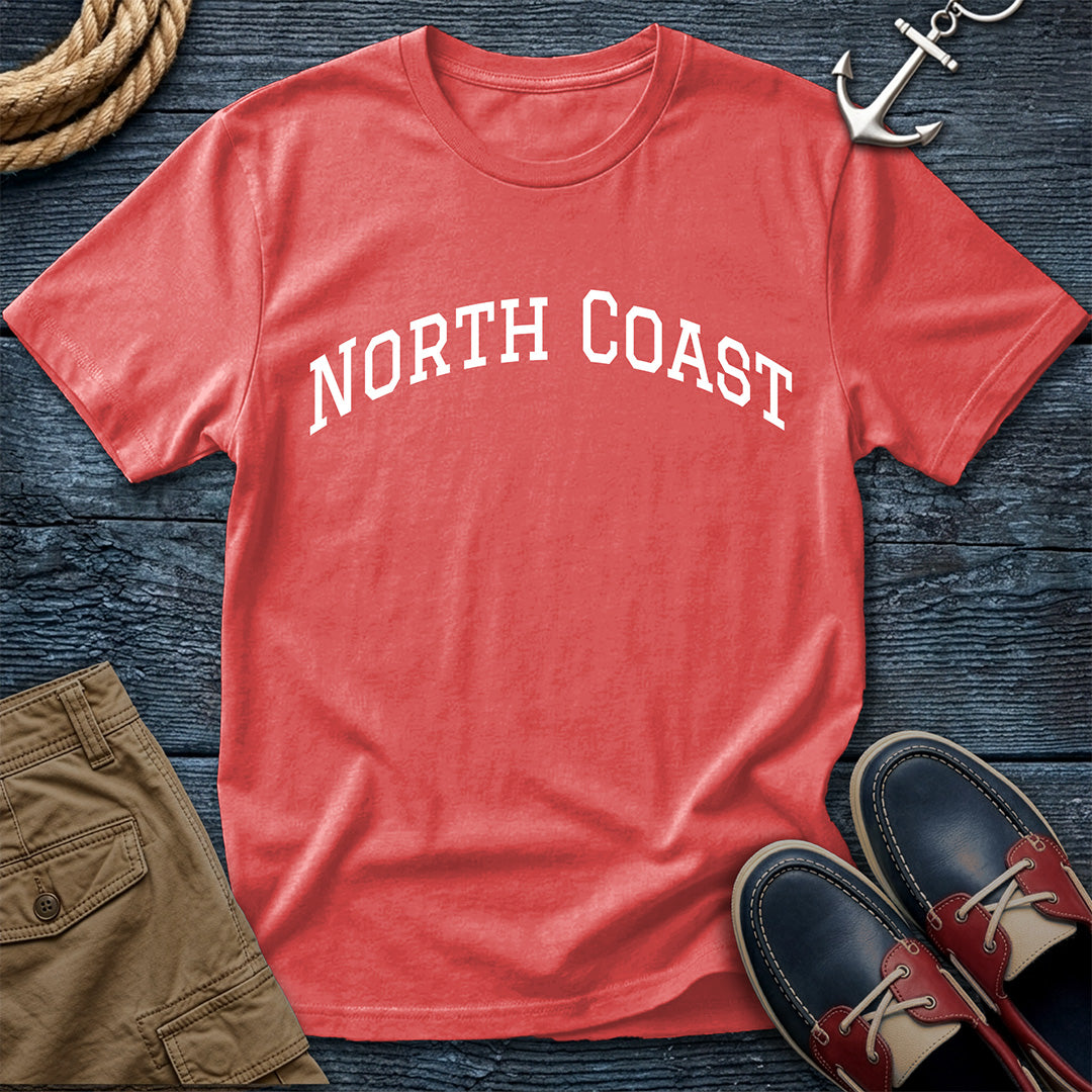 A coral red t-shirt with 'NORTH COAST' printed in white arched text across the chest, styled in a flat lay on a dark wood background with an anchor, boat shoes, shorts, and a rope.