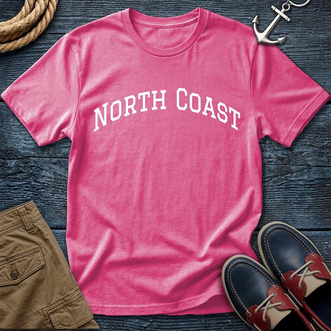 A pink short-sleeve t-shirt with 'NORTH COAST' printed in white arched text across the chest, styled in a flat lay on a dark wood background with a rope, silver anchor, tan shorts, and navy boat shoes.