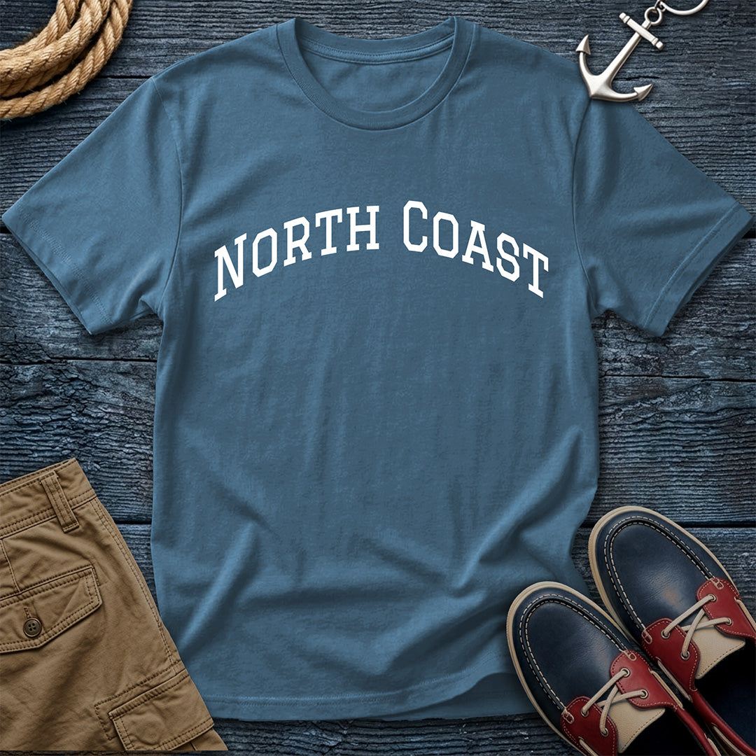 A slate blue crewneck t-shirt with 'NORTH COAST' printed in white arched letters across the chest, styled in a flat lay with brown shorts, red and navy boat shoes, a rope, and an anchor on a dark wood background.