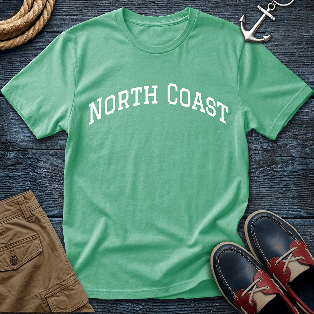 A green short-sleeve t-shirt with 'NORTH COAST' printed in white arched text across the chest, styled in a flat lay on a dark wood background with brown shorts, red and navy boat shoes, a rope coil, and a silver anchor.